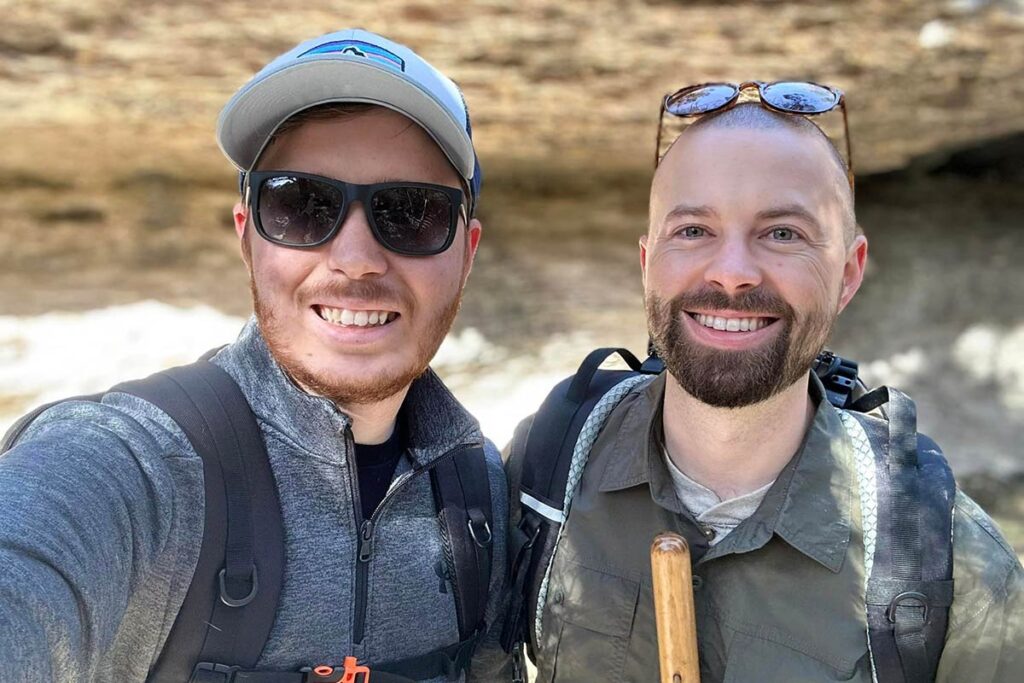 Trenton Ormsbee-Hale on a hike with his husband. Both are wearing backpacks and smiling for the camera.
